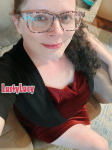 Cam model LustyLucy is looking sexy, holding the phone above to reveal her sitting cross-legged on her boyfriend's chair in a red velvet dress, her long hair pulled to the side and flowing over her shoulder. She is smiling sweetly with her red lipstick lips, her green eyes looking directly at the camera through her big glasses.