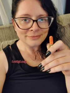 Stoner cam model LustyLucy is wearing a black shirt, with long black fingernails and her long black hair pulled to the side. She is holding up a weed vape, smiling slightly with her happy looking stoned eyes looking at you through her sexy cat eye glasses.