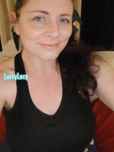 Cam model and content creator LustyLucy smiling with an impish look on her pretty face, her green eyes sparkling. You can see her big chest in her black tank top.